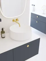 Elegant og moderne baderomsinspirasjon med skråtak, vegghengte blå servantskap og underskap, rund servant med messingarmatur, opplyst speil og naturlig lysinnfall